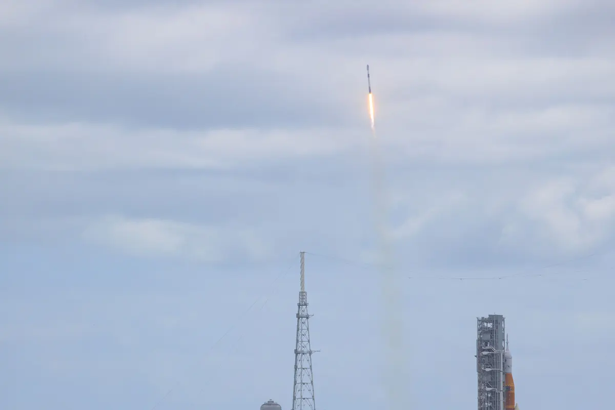 A Falcon 9 rocket climbing into a cloudy sky, with the Artemis I rocket standing on its pad at right